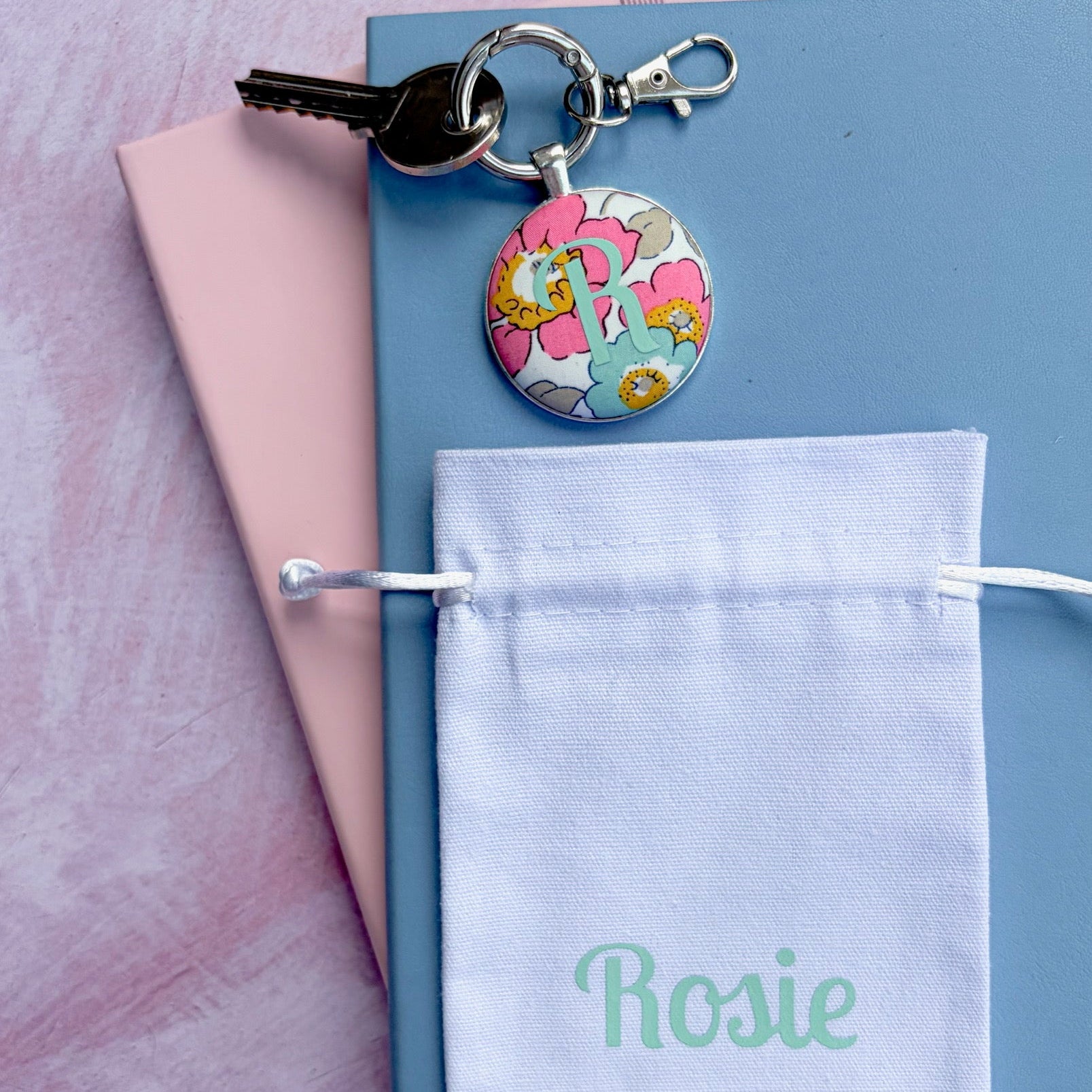 A personalized keyring with a round floral charm and a key, displayed on top of a blue notebook with a pink page, next to a white drawstring pouch printed with the name 'Rosie' in mint green, all on a pink textured background.