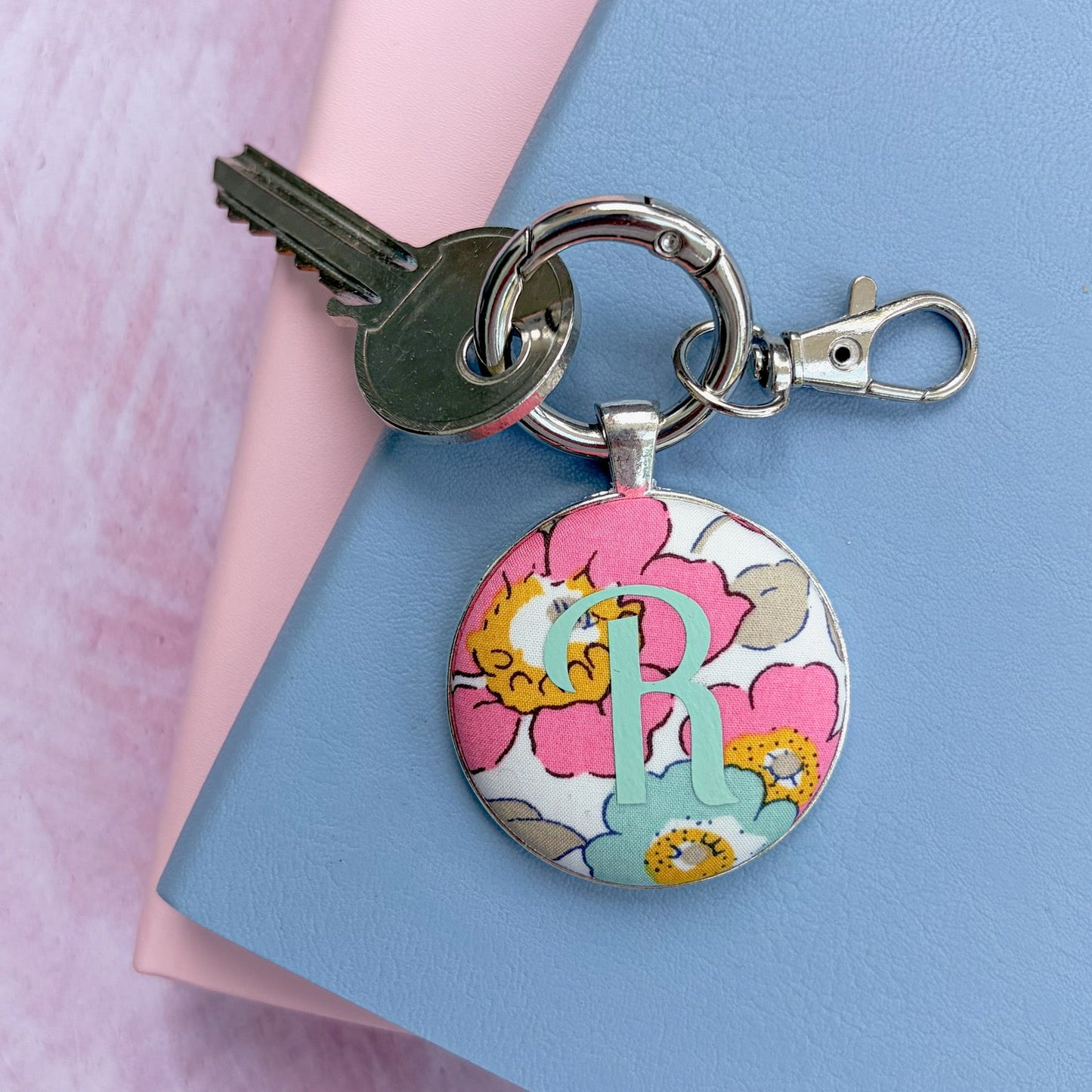 A personalized round keychain with a floral pattern and a monogrammed initial 'R', attached to a keyring with a key and clasp, displayed on pink and blue textured paper.