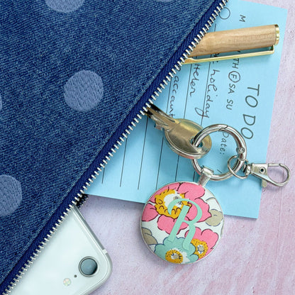 A close-up flat lay of a blue denim pouch with white polka dots, partially unzipped to reveal a light blue notepad, a wooden pen, and a round floral keychain with a silver clasp. A white smartphone is tucked into the pouch, all set on a pink textured surface.