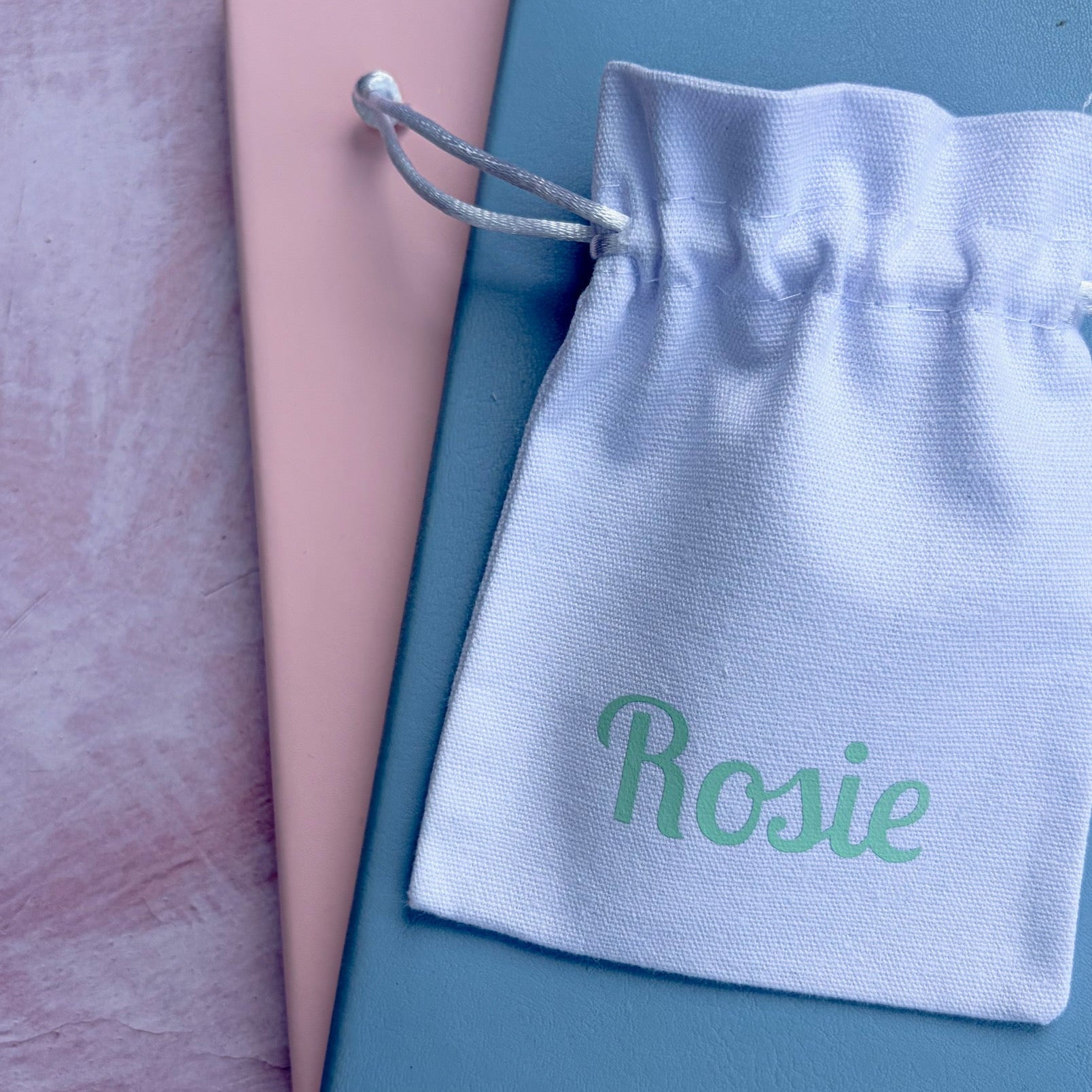 A white drawstring pouch with the name 'Rosie' printed in green, resting on a dusty blue book over a pink book, on a textured pink background.