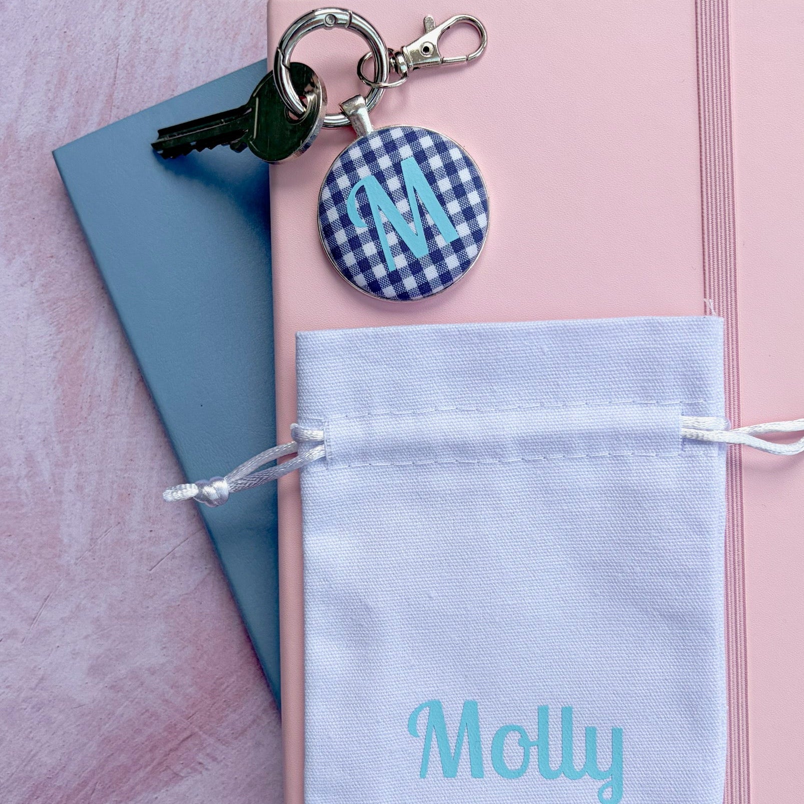 A round personalized keyring made with a navy blue gingham fabric and printed with a turquoise M, on a silver clip ring, with a personalised white drawstring gift pouch with the name Molly in turquoise printed on the front, styled on a pink and blue notebook