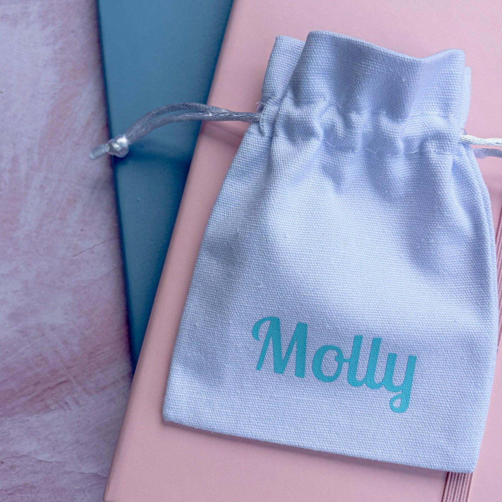 A white personalized drawstring pouch with the name 'Molly' printed in turquoise, resting on top of a pink notebook and a teal book on a textured pink surface.