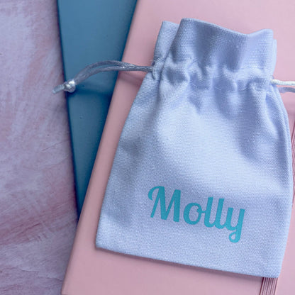 A white personalized drawstring pouch with the name 'Molly' printed in turquoise, resting on top of a pink notebook and a teal book on a textured pink surface.