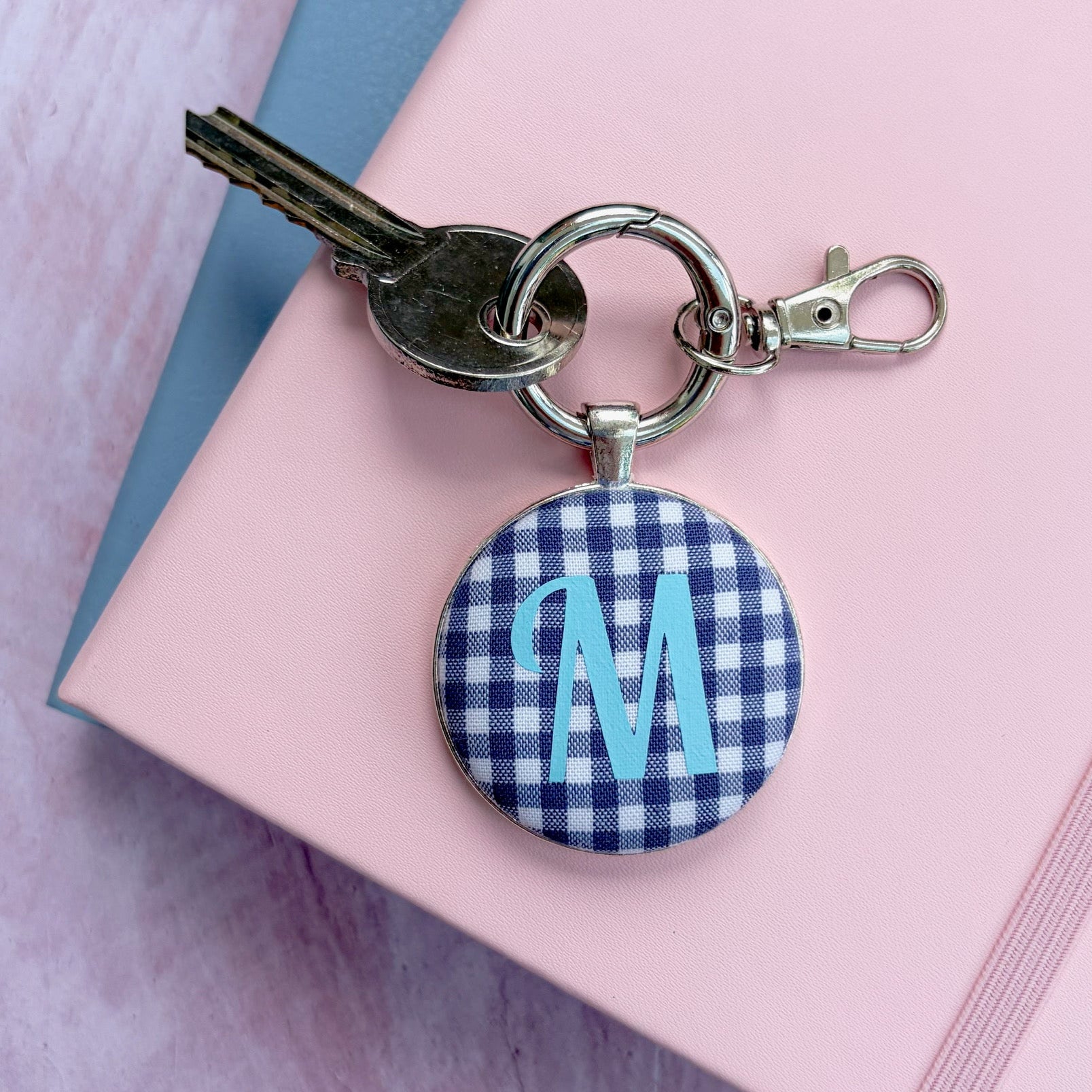 A personalized keychain featuring a round button charm with a navy and white gingham pattern and a light blue monogram letter 'M', attached to a silver keyring with a key and clasp, resting on a pink notebook.