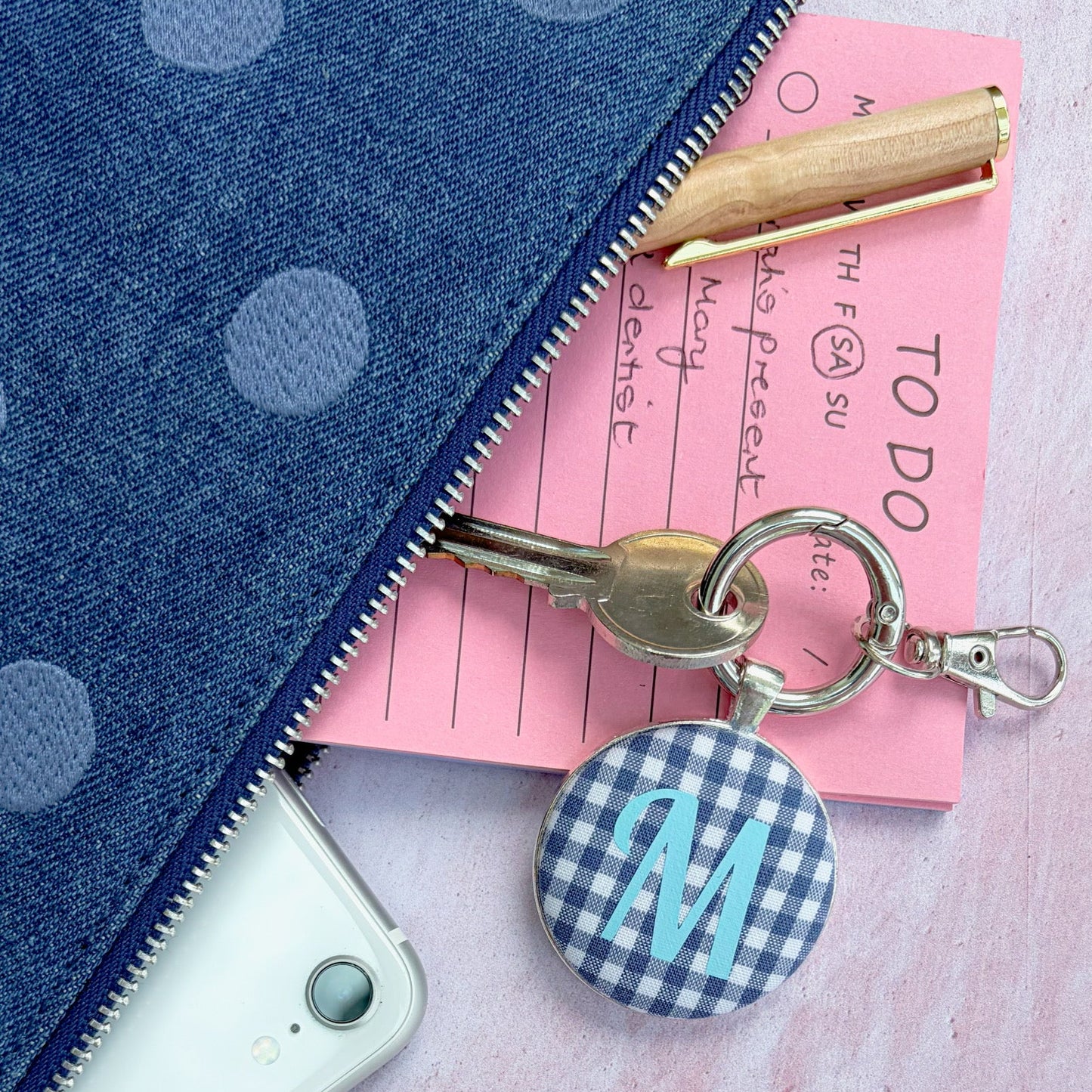 A close-up flat lay of a blue polka dot zipper pouch with a keychain attached. The keychain has a round blue-and-white gingham button with a light blue monogram letter 'M' and a gold keyring.