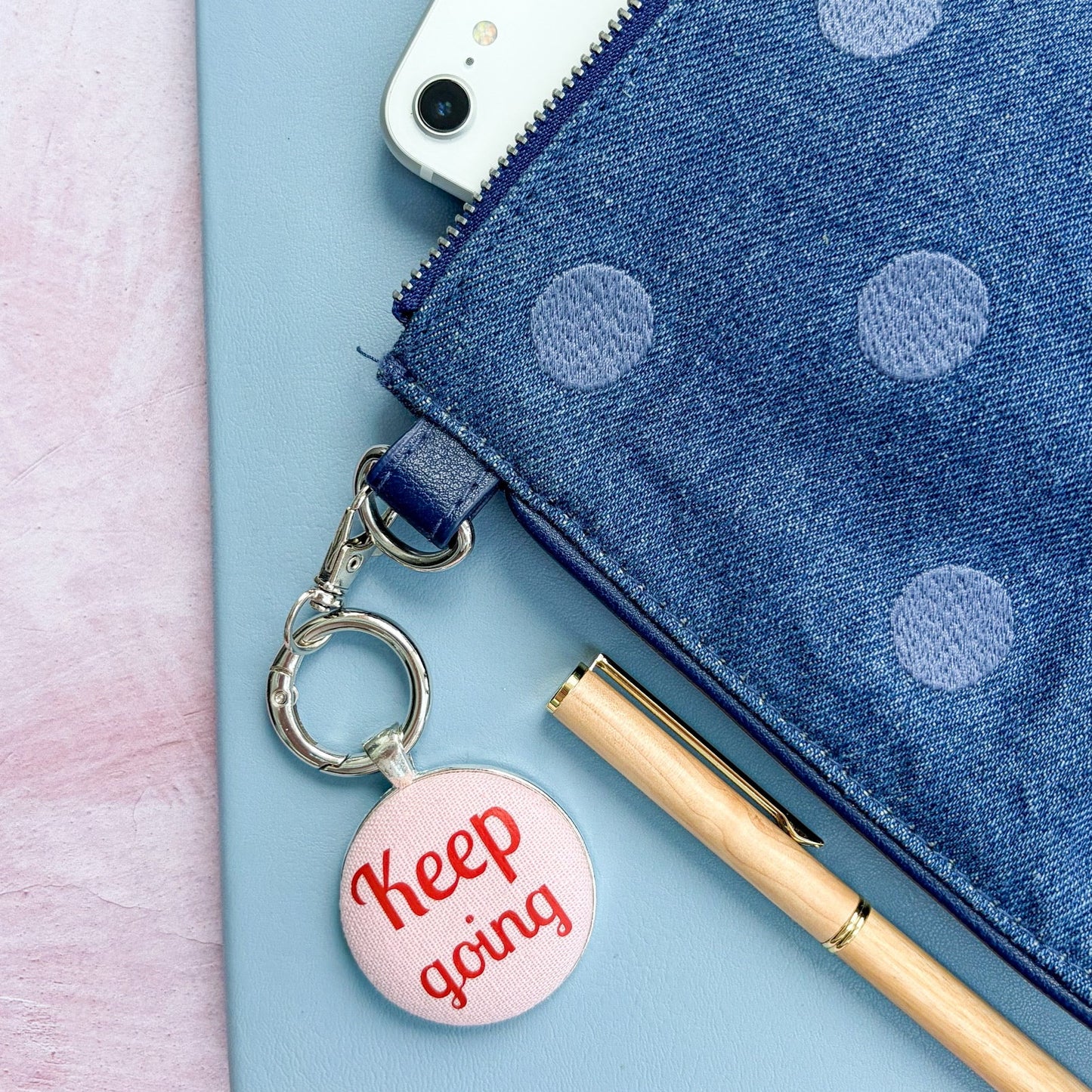 Blue polka dot pouch with a phone, pen, and 'Keep going' keychain on a light pink background