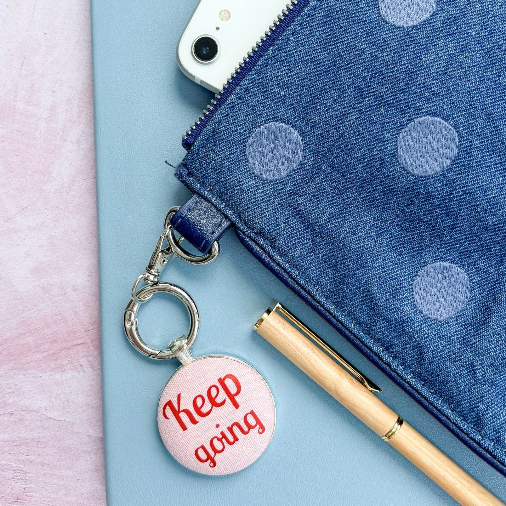 Blue polka dot pouch with a phone, pen, and 'Keep going' keychain on a light pink background