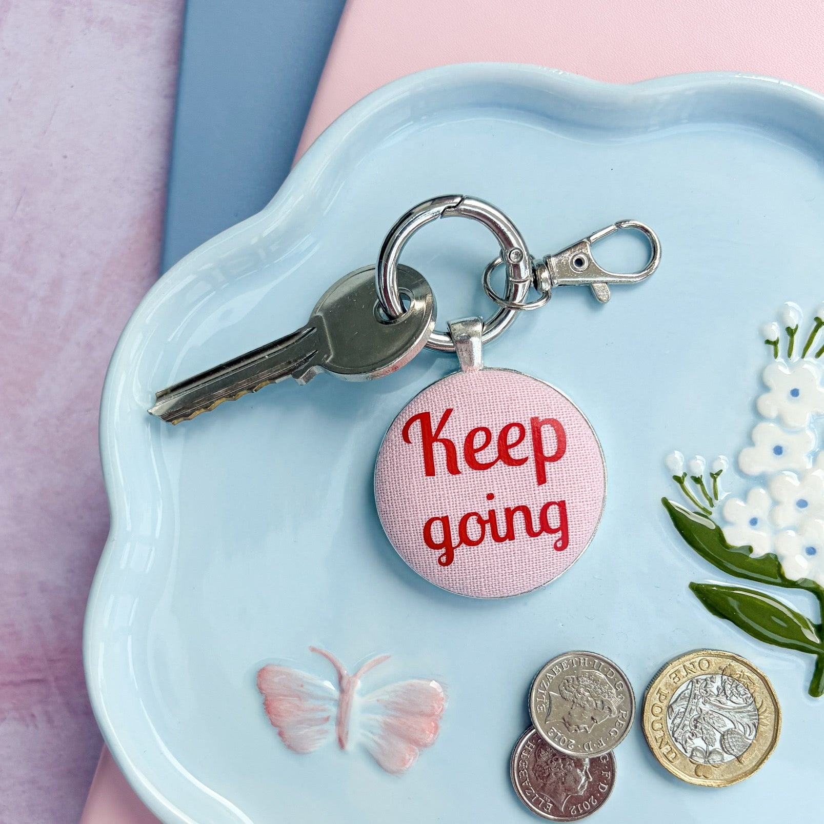 Keyring with 'Keep going' printed in red on pink fabric, on a decorative dish with coins and flowers
