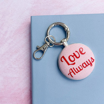 Keyring with 'Love Always' text in red on pink fabric, on a pink and blue background