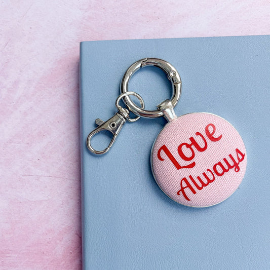 Keyring with 'Love Always' text in red on pink fabric, on a pink and blue background