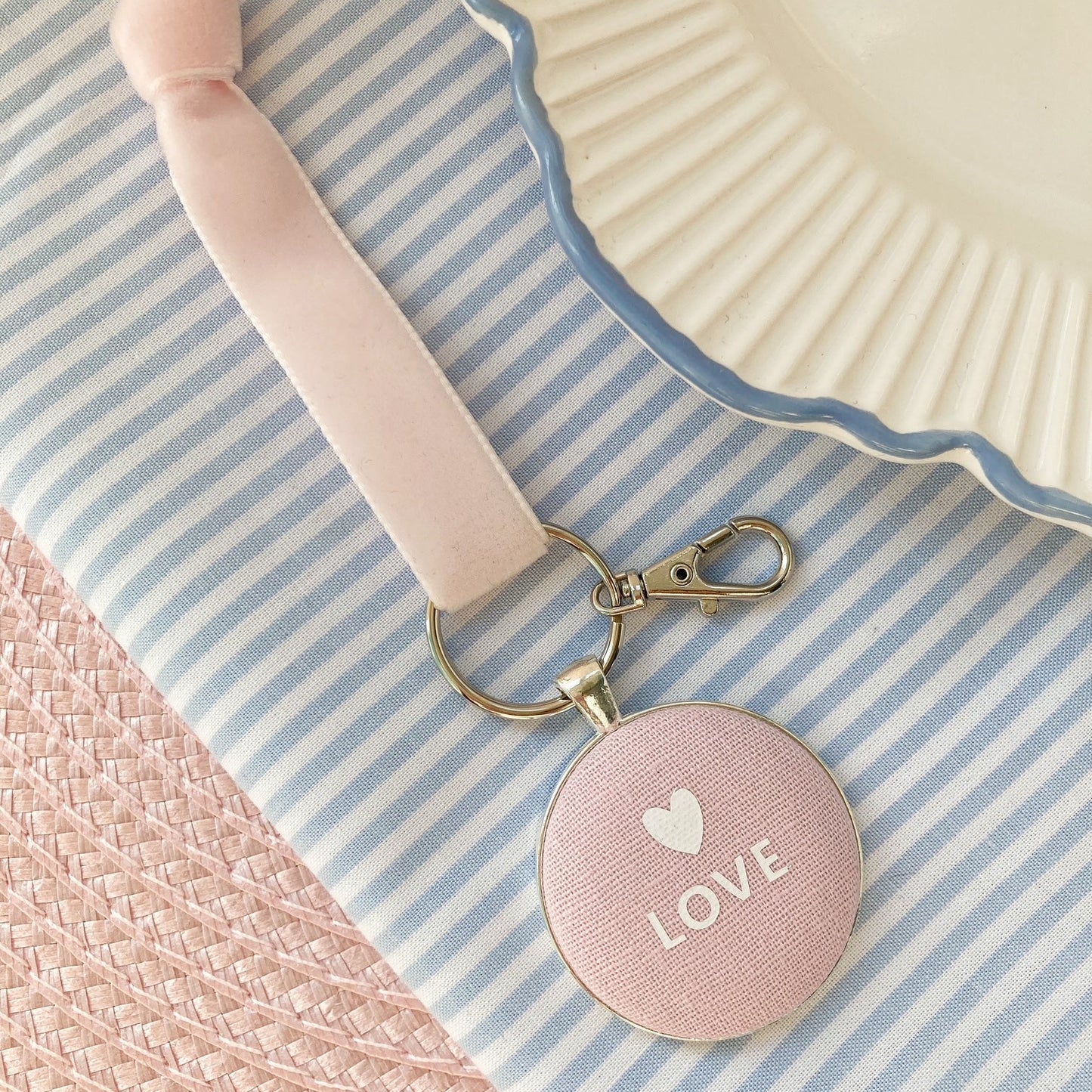 Pink keychain with 'LOVE' and heart symbol on pink fabric, laid on a striped background