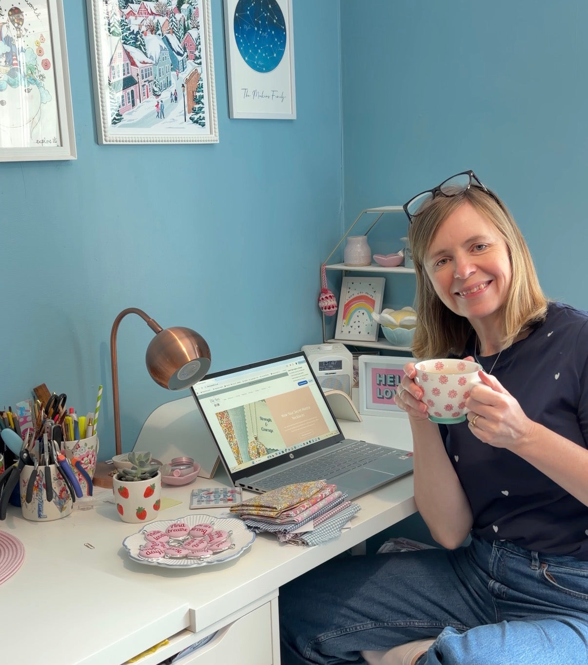 Mandy Meakins, founder of Ellie Bea jewellery sitting at a desk with a laptop and various items, holding a cup.