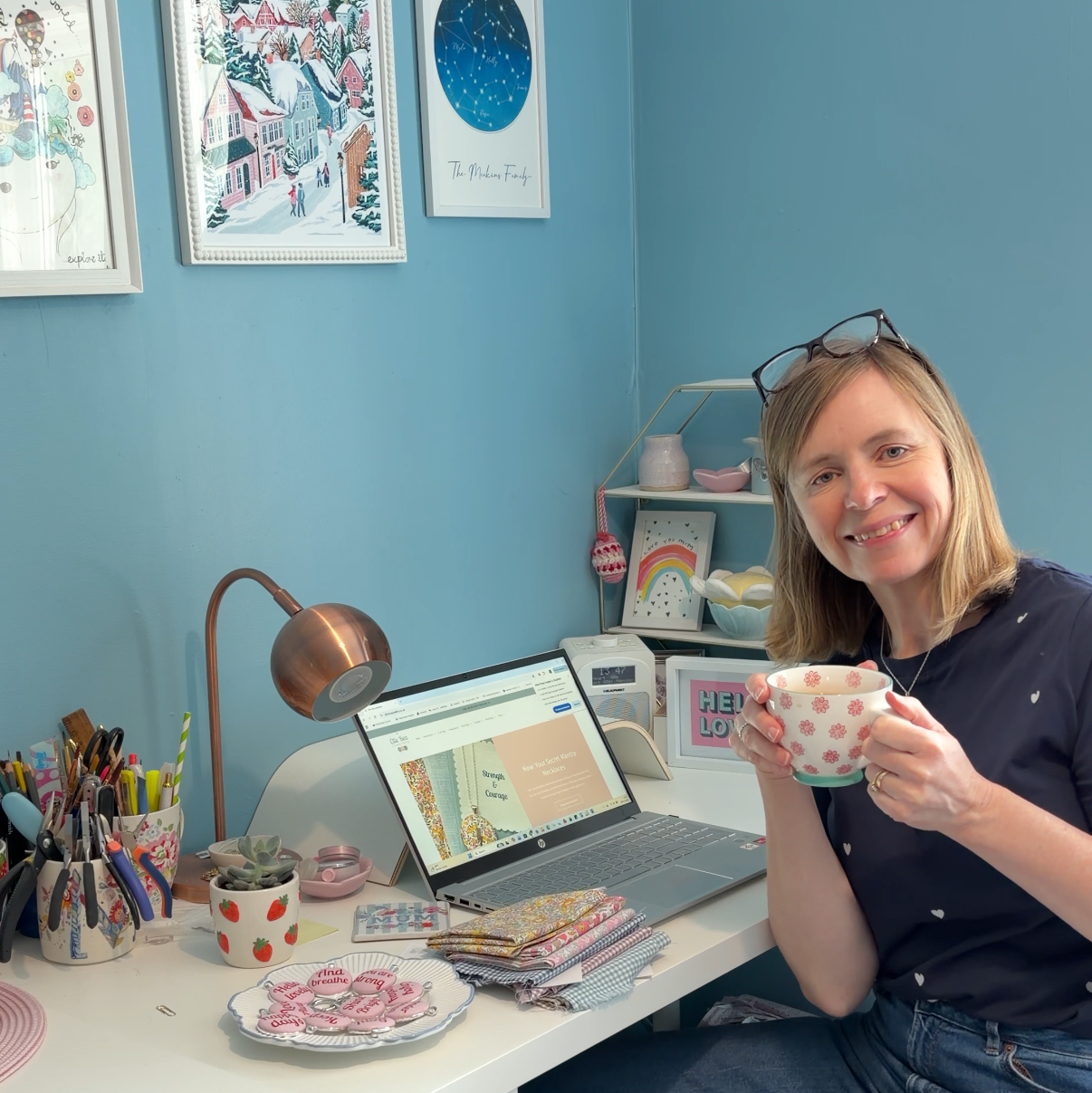 Mandy Meakins, founder of Ellie Bea jewellery sitting at a desk with a laptop and various items, holding a cup.
