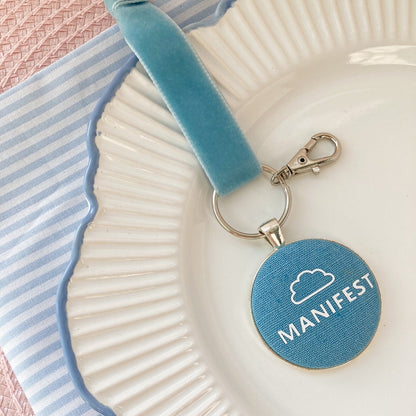 Blue keepsake with 'MANIFEST' and a cloud on blue fabric, laid on a decorative plate