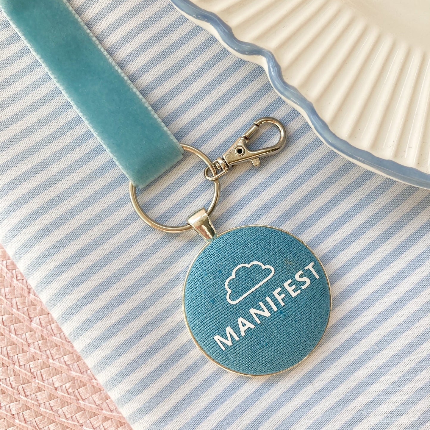 Blue keychain with 'MANIFEST' text and white cloud printed on blue fabric