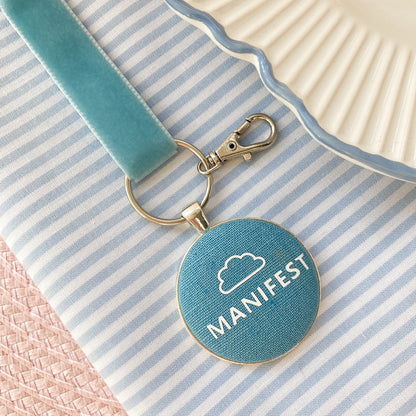 Blue keychain with 'MANIFEST' text and white cloud printed on blue fabric
