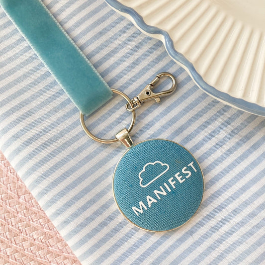 Blue keychain with 'MANIFEST' text and white cloud printed on blue fabric