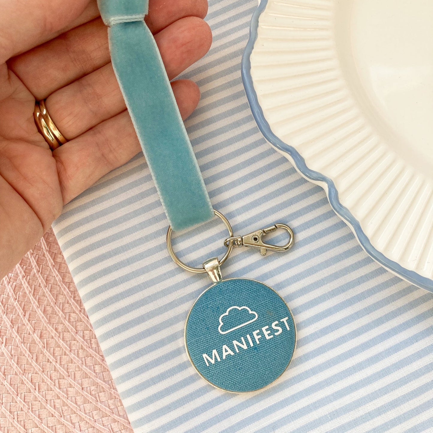 Blue keyring with 'MANIFEST' and cloud icon, on a striped fabric background