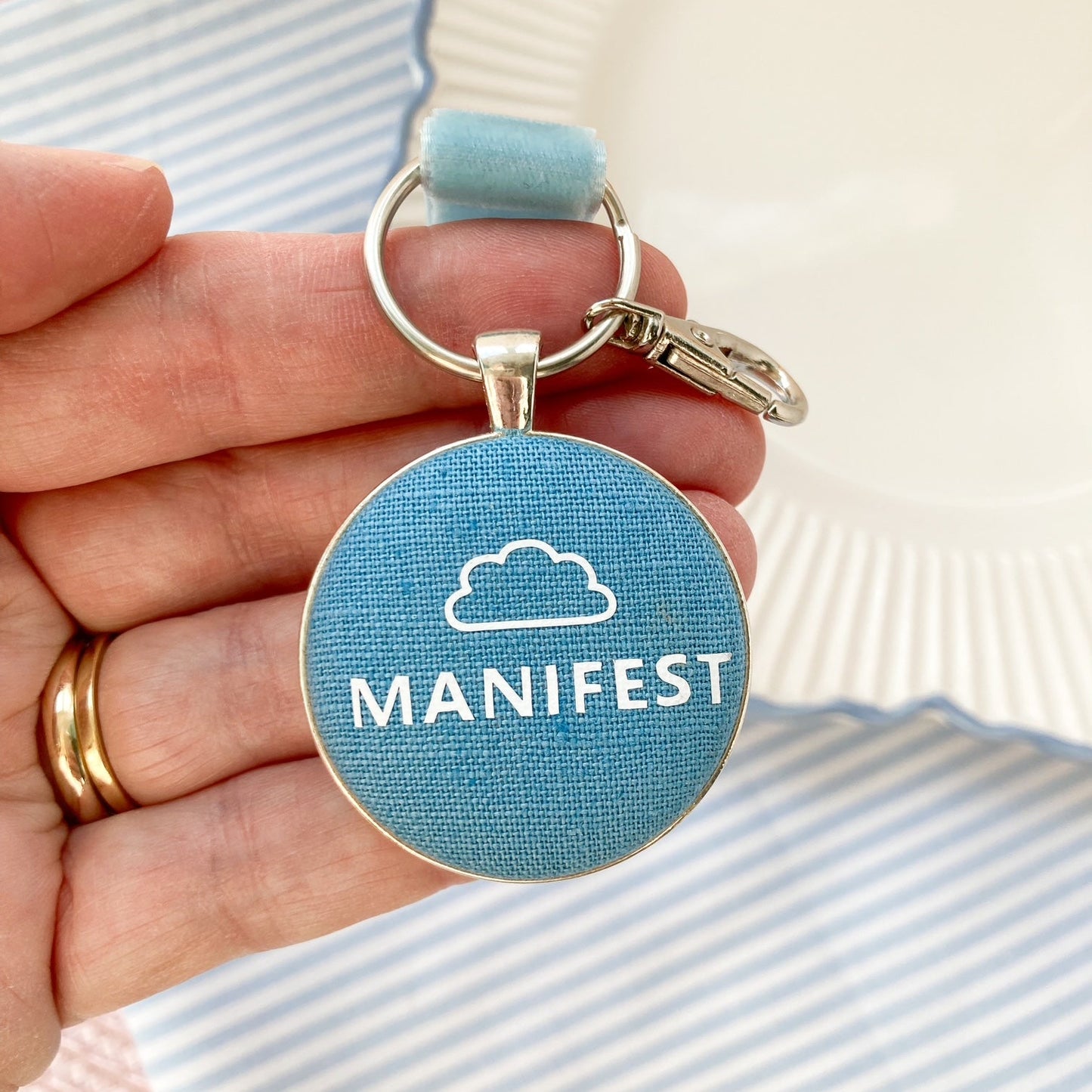 Hand holding a keychain with 'MANIFEST' and cloud logo on a blue background