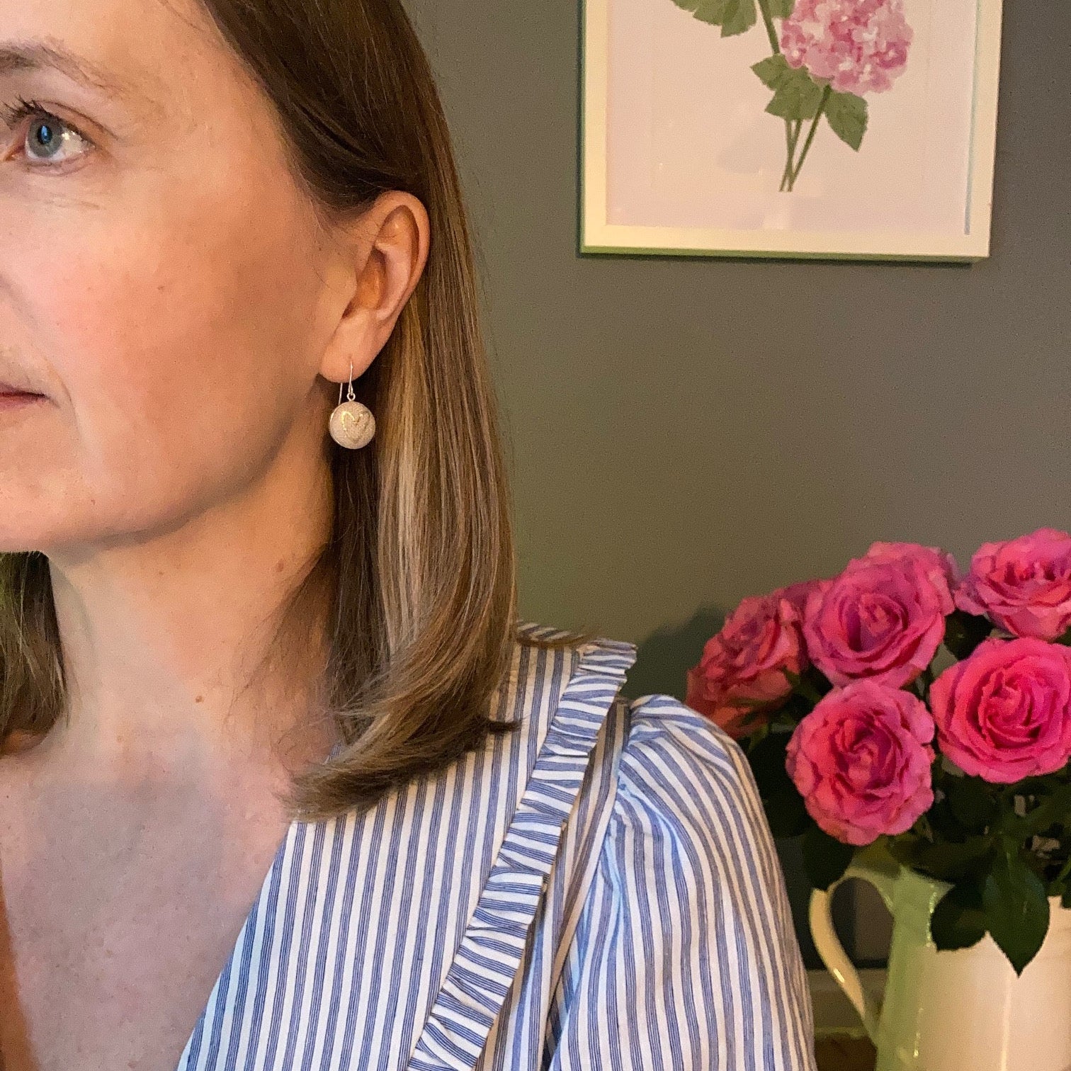 Person wearing a blue and white striped shirt with natural heart earrings with a vase of pink roses in the background.