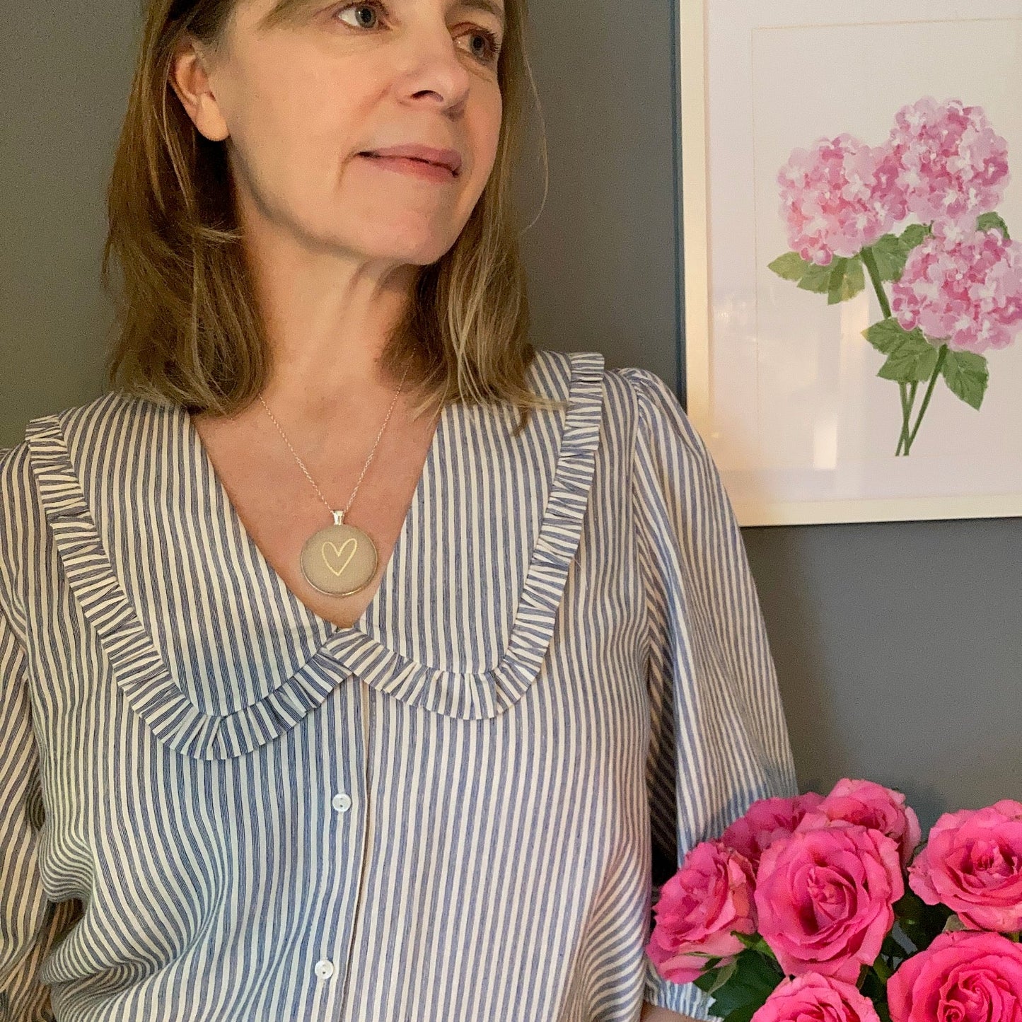 Woman in a striped shirt and natural gold heart necklace holding pink flowers indoors with a floral painting on the wall.
