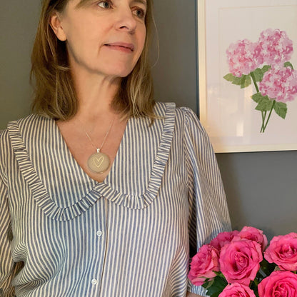 Woman in a striped shirt and natural gold heart necklace holding pink flowers indoors with a floral painting on the wall.