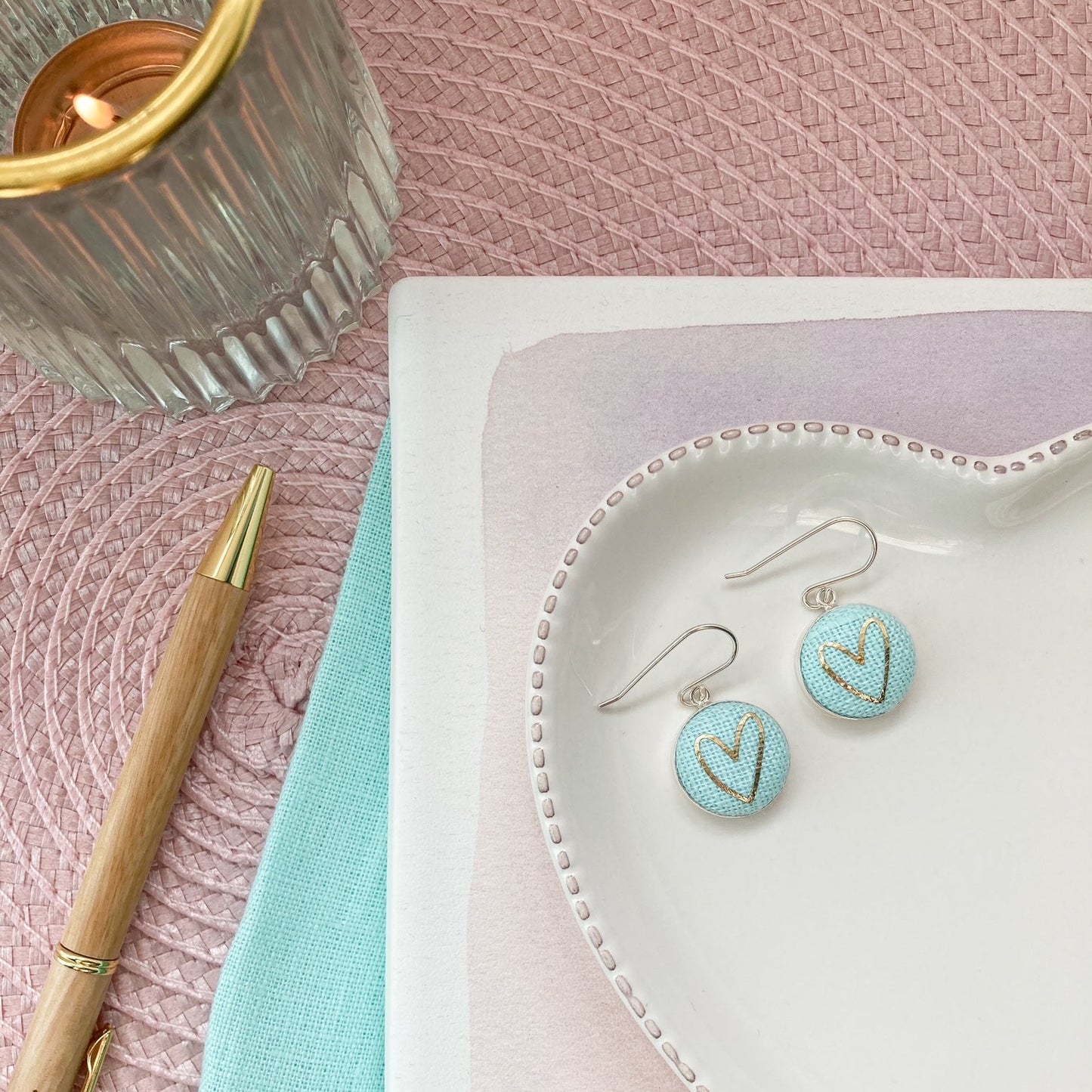 Mint aqua earrings on a heart-shaped white dish with a pen and candle on a pink surface