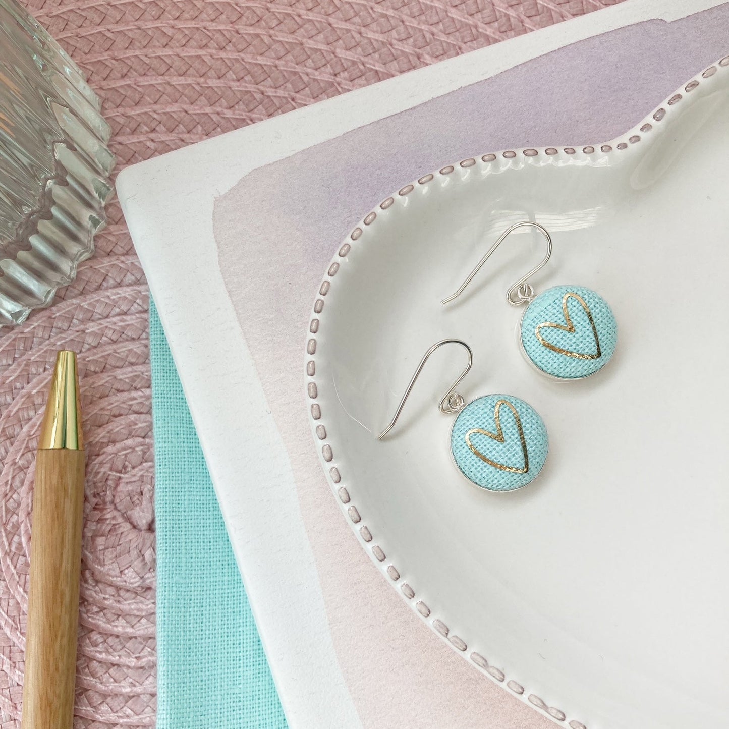 Pair of earrings on a textured pink surface with a pen and notebook.
