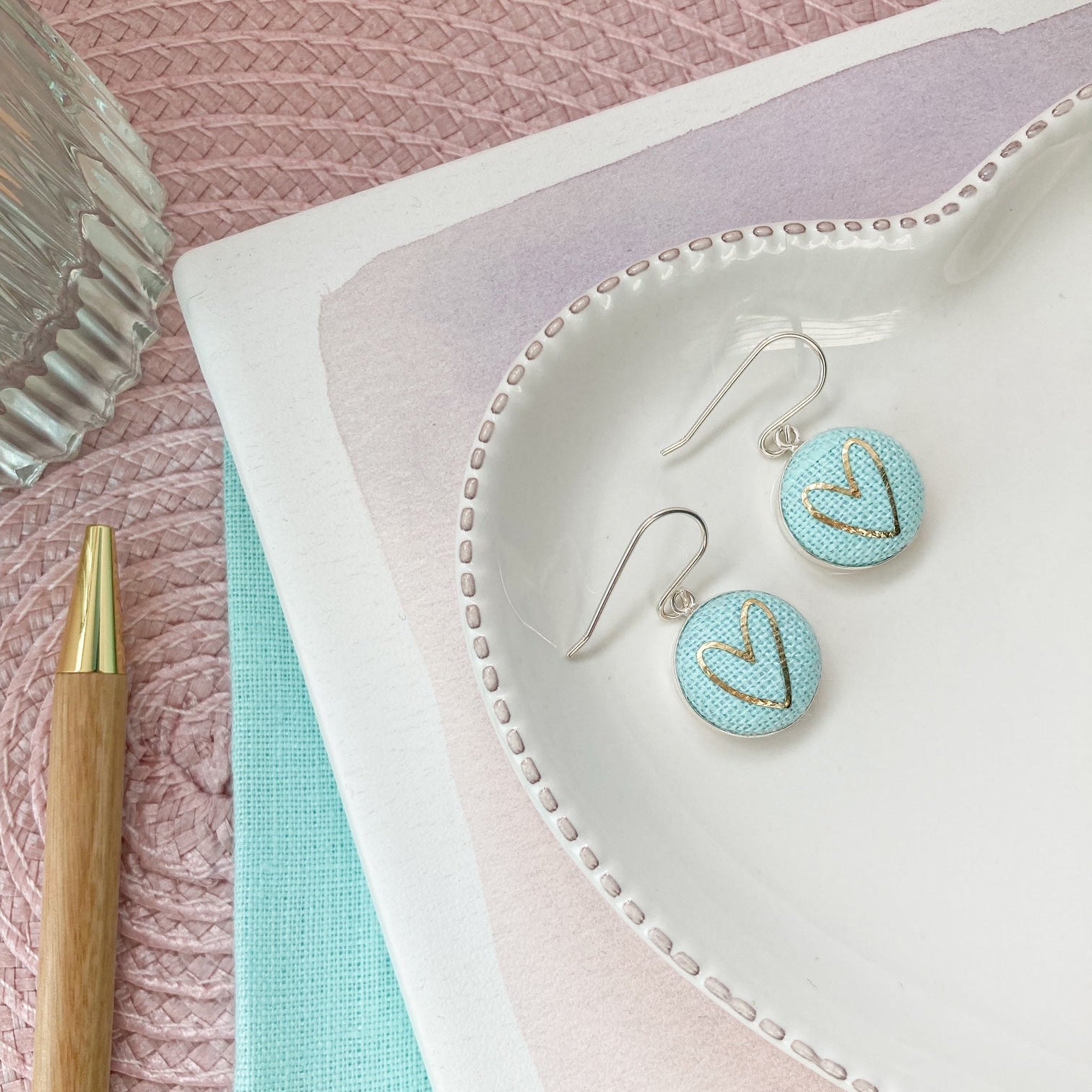 Pair of earrings on a textured pink surface with a pen and notebook.