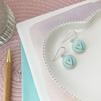 Pair of earrings on a textured pink surface with a pen and notebook.