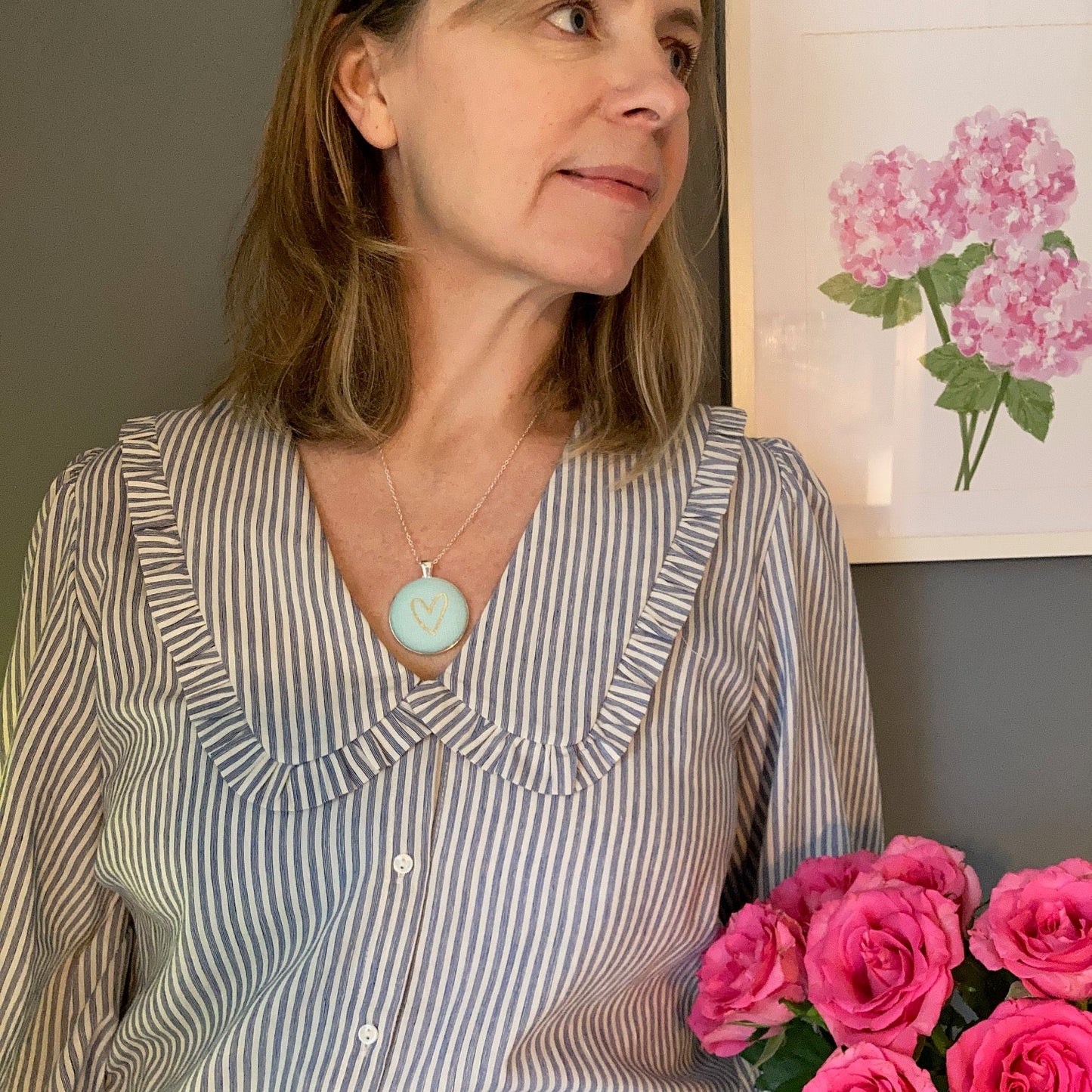 Woman wearing a striped shirt with a aqua heart necklace, holding pink flowers, in a room with a painting of pink flowers on the wall.