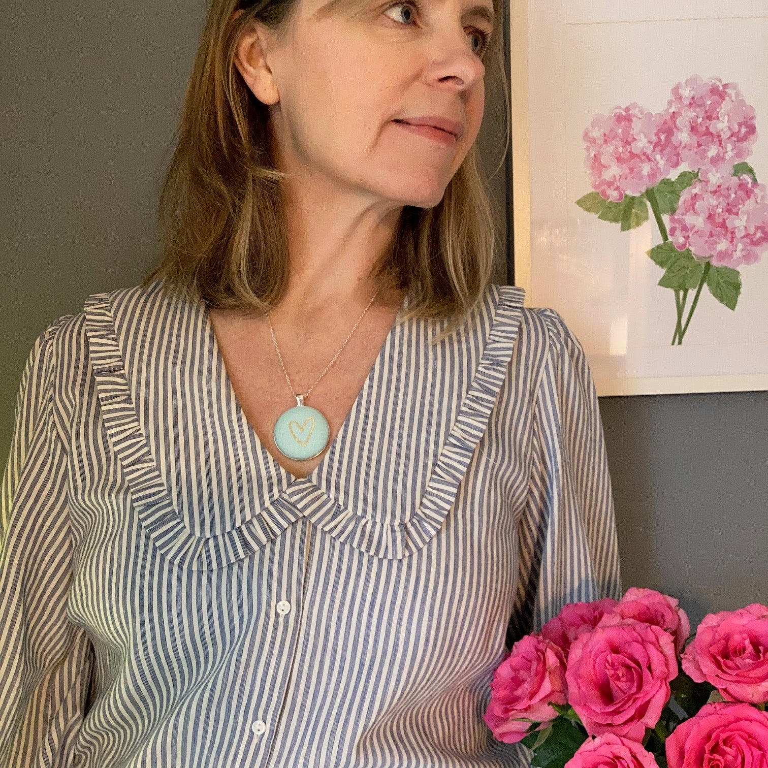 Woman wearing a striped shirt with a aqua heart necklace, holding pink flowers, in a room with a painting of pink flowers on the wall.