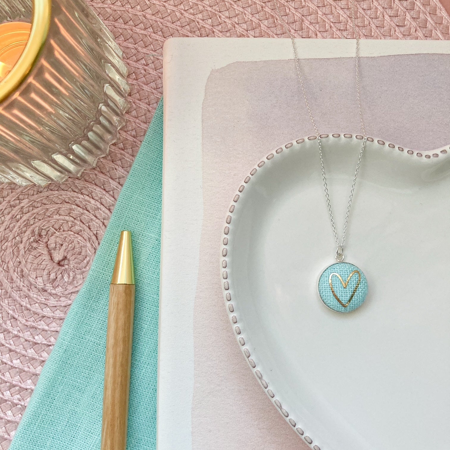Necklace with a heart pendant on a white dish, next to a pen and candle on a textured surface.