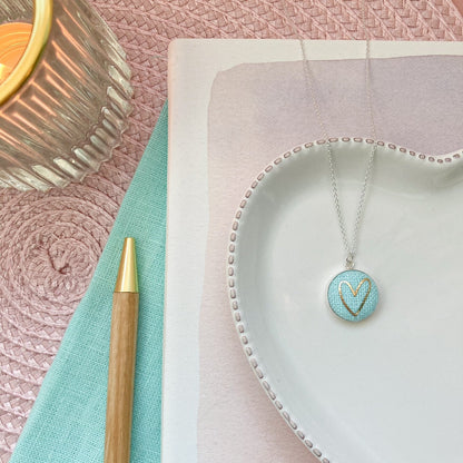 Necklace with a heart pendant on a white dish, next to a pen and candle on a textured surface.