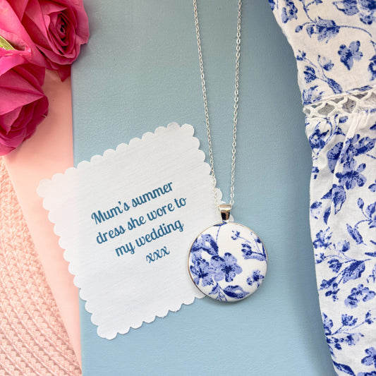 Necklace with blue floral pendant on with a fabric message, surrounded by pink flowers and floral fabric.