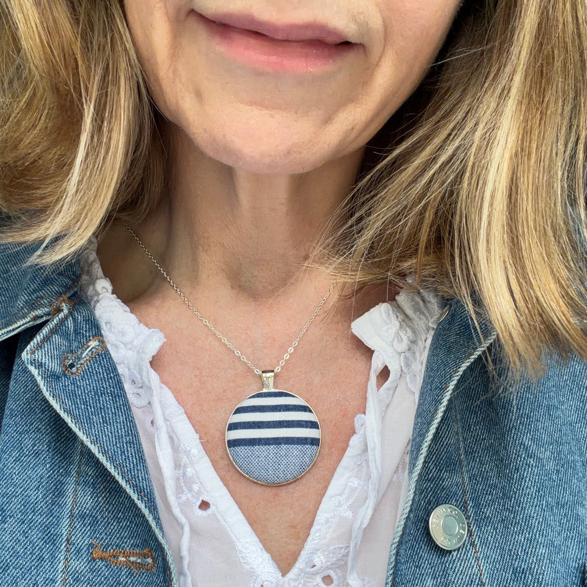 Person wearing a necklace with a circular pendant featuring blue stripes, dressed in a denim jacket and white blouse.