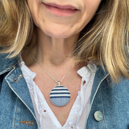 Person wearing a necklace with a circular pendant featuring blue stripes, dressed in a denim jacket and white blouse.