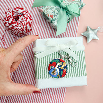 Personalised Christmas Gift Tag and Keyring, made with blue Liberty floral and star fabric, printed with K in gold, set in a keyring, thread on a white velvet ribbon, tied around a small gift and set on a Christmas flatlay