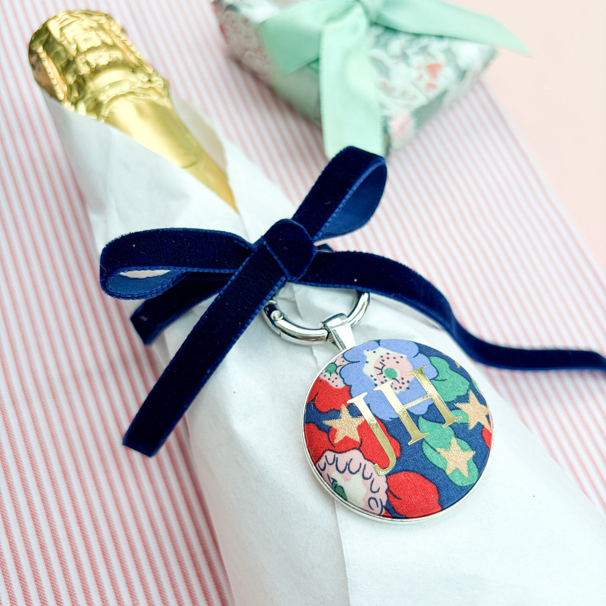 Personalised Christmas Gift Tag and Keyring, made with blue Liberty floral and star fabric, printed with JH in gold foil, set in a keyring, thread on a blue velvet ribbon, tied around a bottle of wine and set on a Christmas red striped fabric