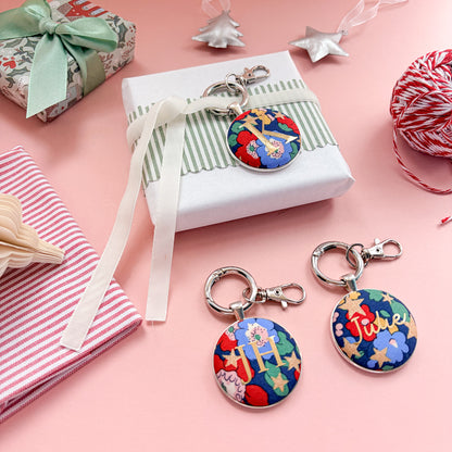 Personalised Christmas Gift Tags and Keyrings, made with blue Liberty floral and star fabric, printed with names and initials in gold, set in a keyring, thread on a white velvet ribbon, tied around a small gift and set on a Christmas flatlay