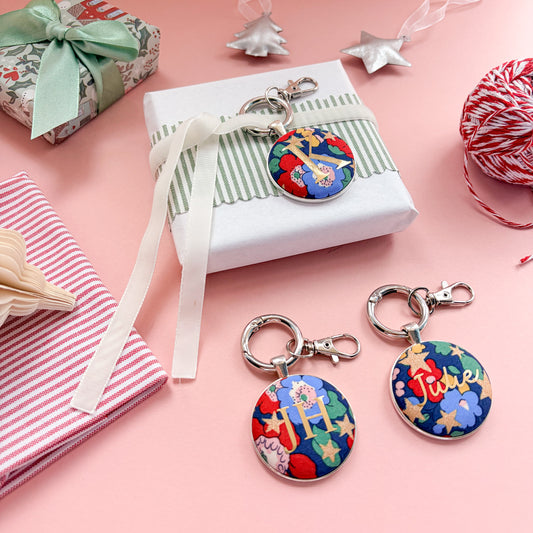 Personalised Christmas Gift Tags and Keyrings, made with blue Liberty floral and star fabric, printed with names and initials in gold, set in a keyring, thread on a white velvet ribbon, tied around a small gift and set on a Christmas flatlay
