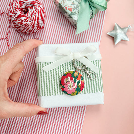 Green floral personalised Christmas gift tag which doubles up as a keyring, made with Liberty fabric, with gold initials NP, thread on a white velvet ribbon, wrapped around a small gift, set on a Christmas flatlay