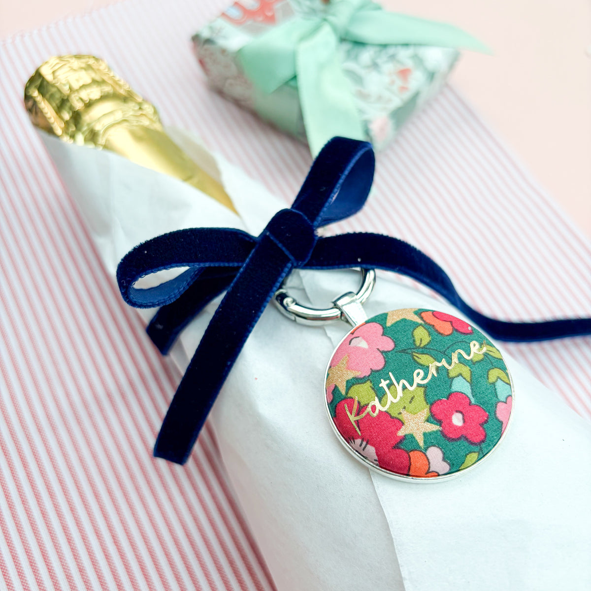 Personalised Christmas Gift Tag Keyring made with green floral Liberty fabric, printed with Katherine in gold, set in a keyring, thread on blue velvet ribbon, tied around a bottle of wine and set on festive red striped fabric