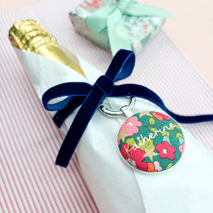 Personalised Christmas Gift Tag Keyring made with green floral Liberty fabric, printed with Katherine in gold, set in a keyring, thread on blue velvet ribbon, tied around a bottle of wine and set on festive red striped fabric