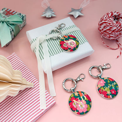 Personalised Christmas gift tags which doubles up as keyrings, with green floral fabric, printed with gold oinitials or name, wrapped around a small gift with white velvet ribbon and set on a Christmas flatlay