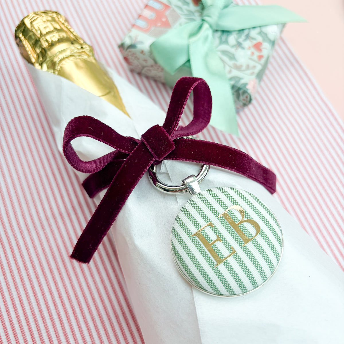 Personalised Christmas Gift Tag and Keyring, made with festive green striped fabric, printed with initials EB in gold foil, set in a keyring, thread on a burgundy velvet ribbon, tied around a bottle of wine and set on a Christmas red striped fabric