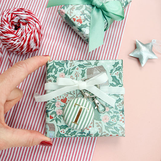 Personalised Christmas Gift Tag and Keyring, made with festive green striped fabric, printed with initial T in gold foil, set in a keyring, thread on a white velvet ribbon, tied around a small gift and set on a Christmas flatlay