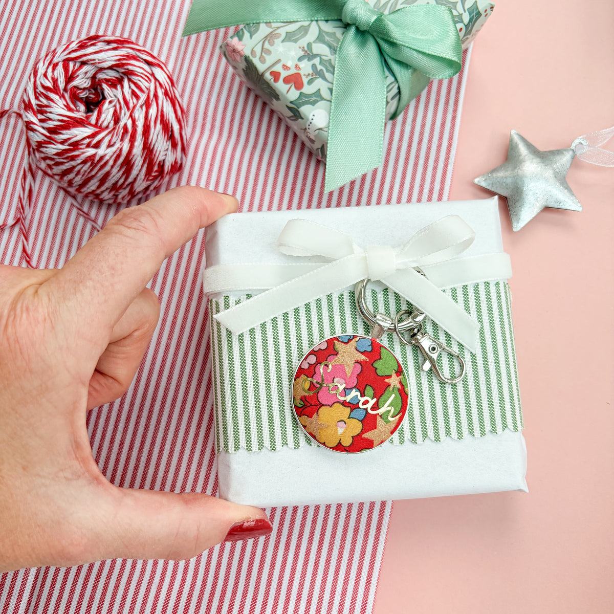 Personalised gift tag which doubles up as a keyring, made with Liberty of London red floral and star fabric, with Sarah in gold print, thread on a white velvet ribbon, wrapped around a small gift, on a Christmas flatlay