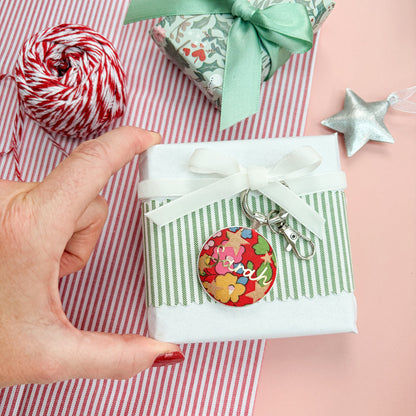 Personalised gift tag which doubles up as a keyring, made with Liberty of London red floral and star fabric, with Sarah in gold print, thread on a white velvet ribbon, wrapped around a small gift, on a Christmas flatlay