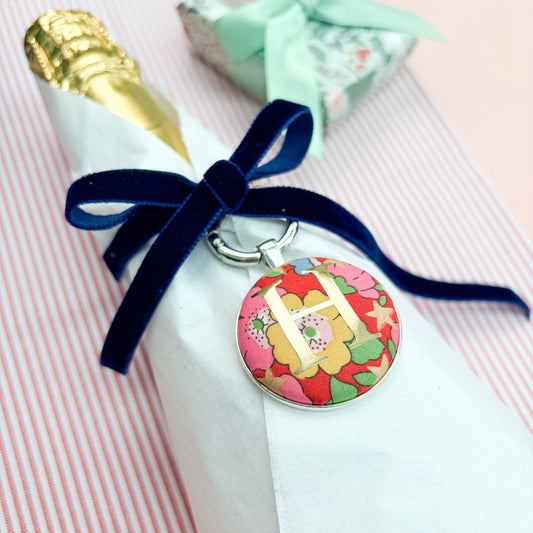 Personalised gift tag which doubles up as a keyring, made with Liberty of London red floral and star fabric, with an H in gold print, thread on a navy blue velvet ribbon, wrapped around a bottle of wine, set on red striped fabric